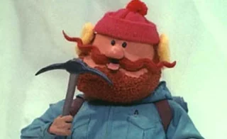 Yukon Cornelius from Rudolph the Red-Nosed Reindeer holding his pickaxe, smiling with his signature red beard, winter hat, and blue coat.